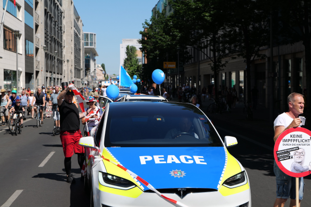 Gruppe von Menschen, die Fahrräder auf einer Straße fahren, mit einem Polizeiwagen im Vordergrund, Bäumen und Gebäuden im Hintergrund unter einem klaren blauen Himmel.