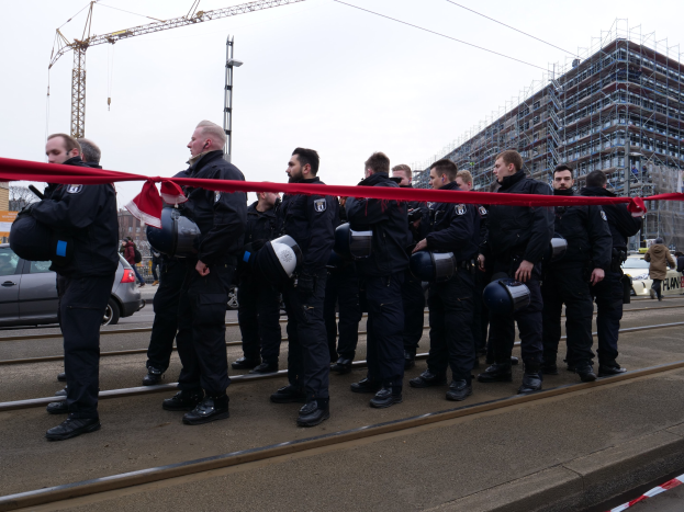 Eine Gruppe von Polizisten in schwarzen Uniformen und Helmen steht in einer Reihe auf einem Bahngleis, mit einem roten Band davor, vor einer Kulisse aus Fahrzeugen, Fußgängern, Gebäuden und einem klaren blauen Himmel.
