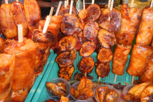 Ein Marktstand mit Spießen von Hähnchenflügeln, verschiedenen Lebensmitteln in Plastikbehältern, mit Menschen und Flaschen im Hintergrund.