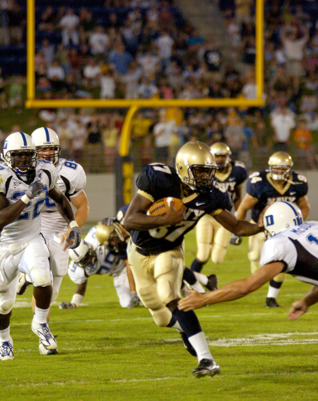 Ein Füßballspieler in einem Sportsdress und Helm rennt mit dem Ball, umgeben von anderen Spielern, mit unscharfem Hintergrund und Text unten.