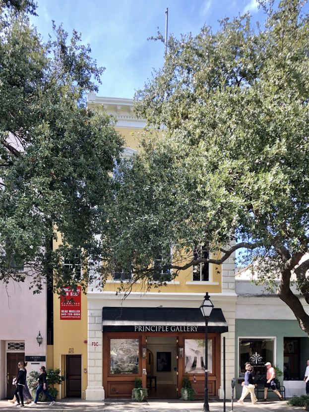 Außenansicht der Principle Gallery in Charleston, SC, mit Menschen, die auf dem Bürgersteig gehen, Topfpflanzen, einer Straßenlaterne, einer Schautafel, einer Fahne, einem Fahnenmast und einem bewölkten Himmel.