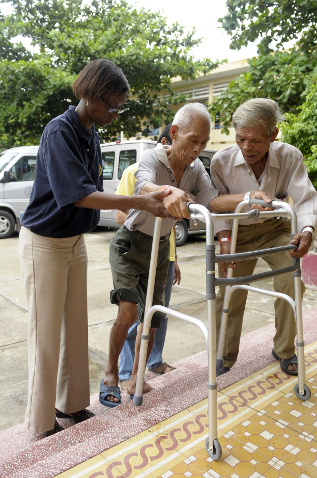 Ein älterer Mann mit einem Rollator wird von einer Krankenschwester übernommen, mit Fahrzeugen, Bäumen, einem Gebäude und einem klaren blauen Himmel im Hintergrund.