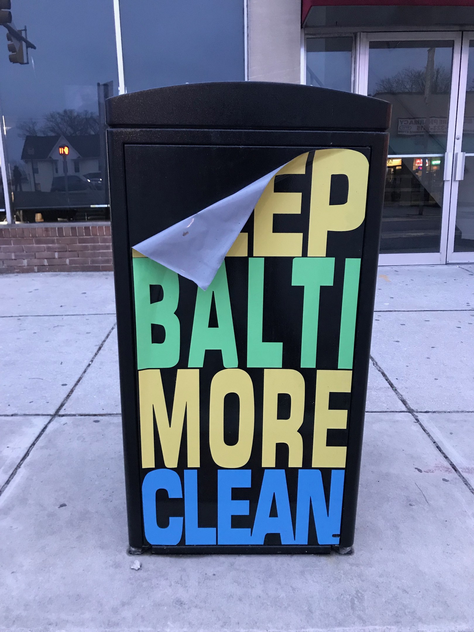 Schwarzer Müllcontainer mit "Keep Baltimore More Clean"-Text auf einem Gehweg vor einem Gebäude mit Glastüren.