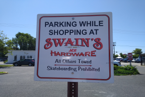 Ein Parkplatz mit einem Schild "Parken beim Einkaufen bei Swain's Hardware" im Vordergrund, mit Fahrzeugen, Pflanzen, Bäumen, Gebäuden, Pfählen, Drähten und einem klaren blauen Himmel im Hintergrund.