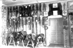 Schwarzes und weißes Foto eines Raums mit verschiedenen Reitzubehör-Artikeln, Fotorahmen und einer Tür an der Wand.