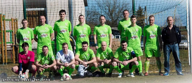 Eine Gruppe von Männern in grünen T-Shirts steht auf einem Fußballfeld, einige in Hockstellung, mit einem Ball vor ihnen in der Nähe eines Torpfostens, umgeben von Bäumen, Gebäuden und Fahrzeugen unter einem Himmel.