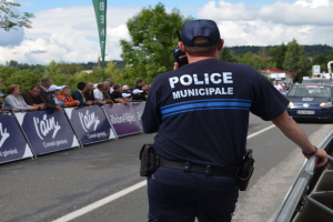 Polizist in Uniform mit Mütze und Funkgerät vor einer Menge mit Transparenten, Fahrzeugen, Bäumen, Gebäuden und einem bewölkten Himmel im Hintergrund.
