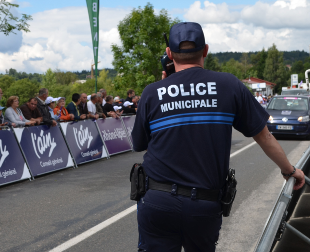 Polizist in Uniform mit Mütze und Funkgerät vor einer Menge mit Transparenten, Fahrzeugen, Bäumen, Gebäuden und einem bewölkten Himmel im Hintergrund.