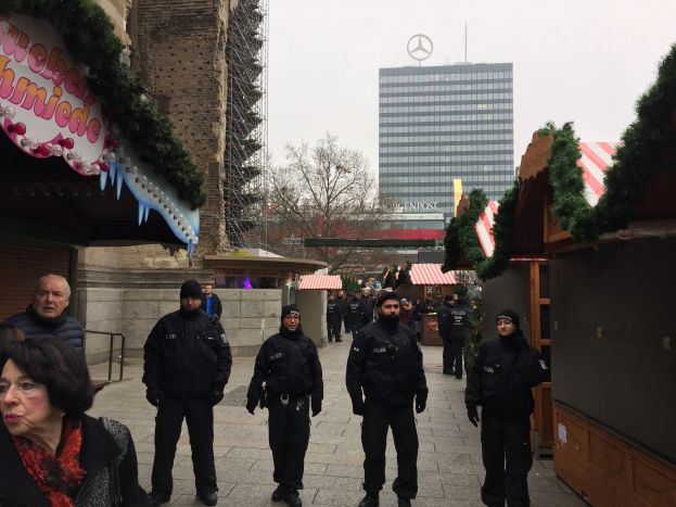 Polizeibeamte in schwarzen Uniformen stehen vor einem geschmückten Weihnachtsmarkt in Berlin mit festlichen Deko, Gebäuden, Bäumen und einem klaren blauen Himmel im Hintergrund.