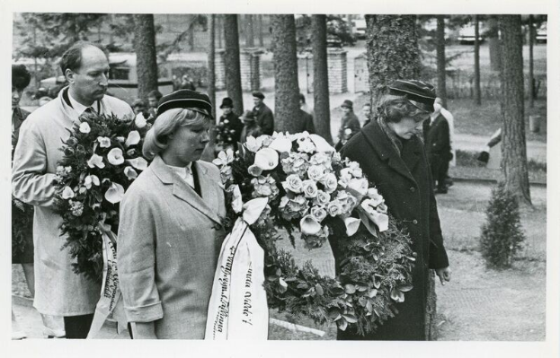 Schwarz-weißes Bild einer Gruppe von Menschen, die Blumensträuße an einem Gedenkdienst für Holocaust-Opfer halten, mit Bäumen und Gebäuden im Hintergrund.