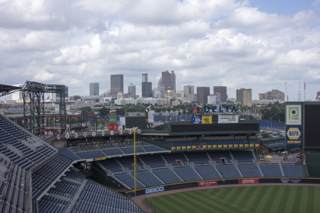 Baseballstadion mit einer Stadtkulisse im Hintergrund, gefüllt mit Stühlen, Pfosten, Brettern und anderen Gegenständen, grasbewachsenem Boden, Bäumen, Gebäuden in der Ferne und einem bewölkten Himmel.