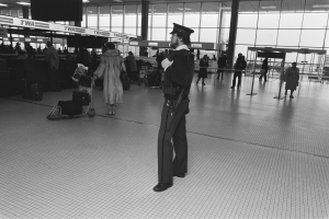 Polizeibeamter in Uniform mit Waffe in einem Flughafen-Terminal umgeben von Menschen und Gepäck.