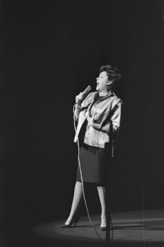 Eine Frau in einem schwarzen und weißen Outfit aus den 1950er-Jahren singt fröhlich auf einer von einem Spotlight beleuchteten Bühne und hält ein Mikrofon.