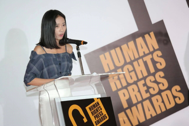 Eine Frau in einem schwarzen Kleid mit einem Dutt steht an einem Rednerpult mit einem Mikrofon und hält eine Rede oder Präsentation vor einem Banner mit der Aufschrift 'Human Rights Press Awards.'