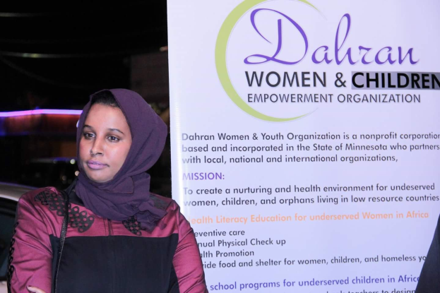 Eine Frau in einem weißen Hemd und blauen Jeans steht entschlossen vor einer Banner für "Dahran Women & Children Empowerment Organization", mit einem Auto im Hintergrund.