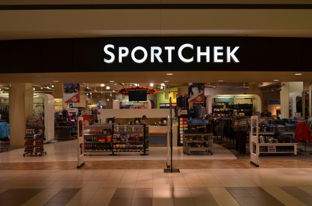 Außen- und Innenansicht eines Sportchek-Verkaufsgeschäfts in einem Einkaufszentrum, mit Kleiderstangen, Deckenleuchten, Säulen und einem Fernseher.