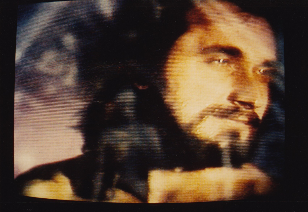 Ein Mann mit Bart und Schnurrbart auf einem Fernseher, der ein weißes Hemd trägt und einen ernsten Gesichtsausdruck hat.