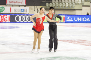 Zwei Eiskunstläufer, ein Mann und eine Frau, gleiten Hand in Hand über eine Eisbahn, umgeben von Zuschauern und Event-Aufbau.