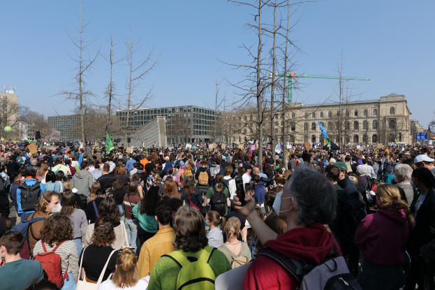 Eine große Menschenmenge bei einer Klimaprotestaktion vor einem Gebäude mit Bäumen und einem klaren Himmel im Hintergrund.