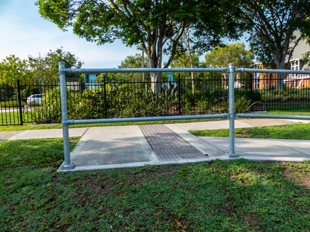 Metallzaun in einer grasbewachsenen Area mit Bäumen und Pflanzen, begrenzt von einer Straße mit einem Auto, Gebäude im Hintergrund und einem klaren blauen Himmel.