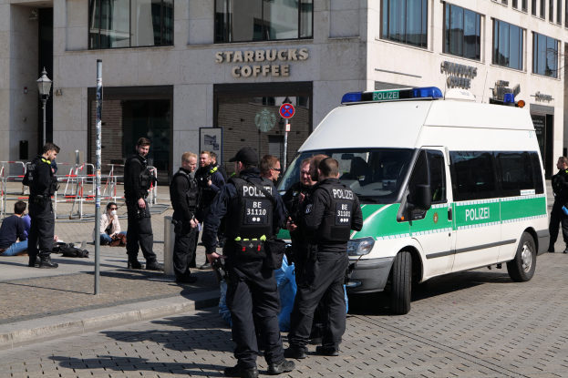 Eine Gruppe von Polizisten steht vor einem Starbucks-Kaffeehaus, mit einem Van auf der rechten Seite und einigen Menschen auf der linken Seite, vor einem Hintergrund aus Gebäude, Schilder, Laterne und Zaun.