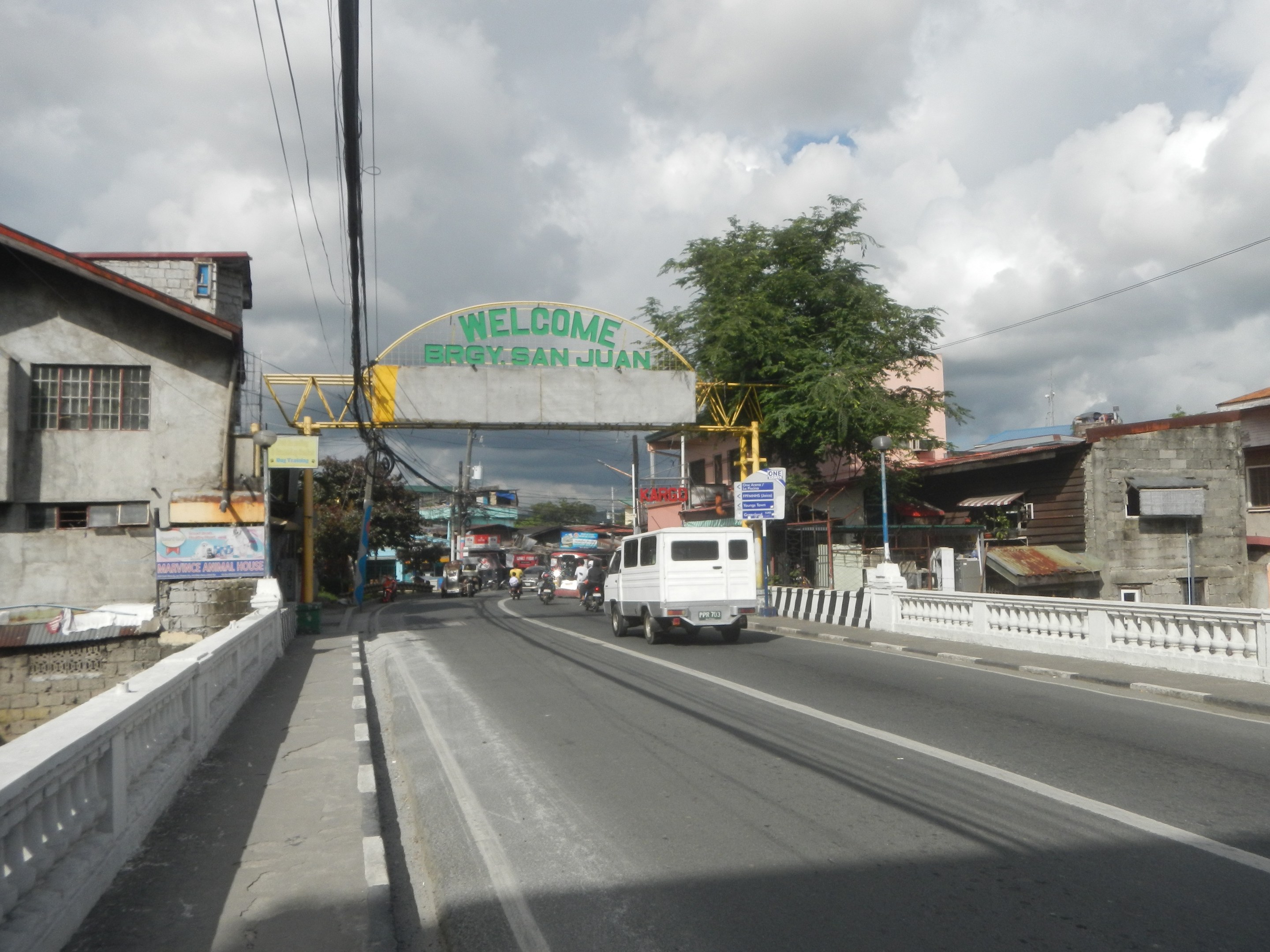 Stadtstraße mit Fahrzeugen, einer Brücke, einem Bogen mit Text, Gebäuden, Bäumen, Strommasten mit Drähten und einem bewölkten Himmel.