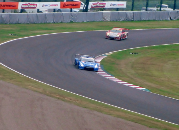 Zwei Autos auf einer Rennstrecke mit Gras auf beiden Seiten, umgeben von Bannern, Maschendrahtzaun, Pfosten und Bäumen.