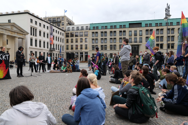 Eine Gruppe von Menschen, die auf dem Boden vor einer Menge sitzt, die Fahnen und Transparente hält, während einer Demonstration gegen Homosexuelle in Berlin, mit einer Statue, einem Mikrofonständer und Gebäuden im Hintergrund.