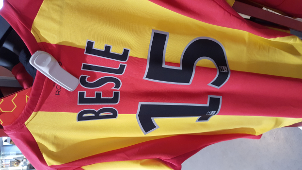 Roter und gelber Fußballtrikot mit aufgedrucktem Text und Nummern, das an einer Ladengestell hängt, mit anderen Gegenständen und dem Boden im Hintergrund.