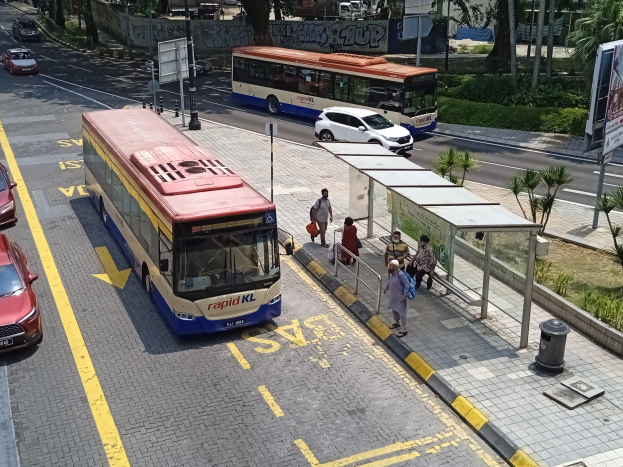 Stadtstraße mit fahrenden Autos und Bussen, eine Bushaltestelle auf dem Bürgersteig mit Fußgängern und umliegende städtische Elemente einschließlich Pfosten, Schildern, Bäumen, Pflanzen, Gras und Gebäuden.
