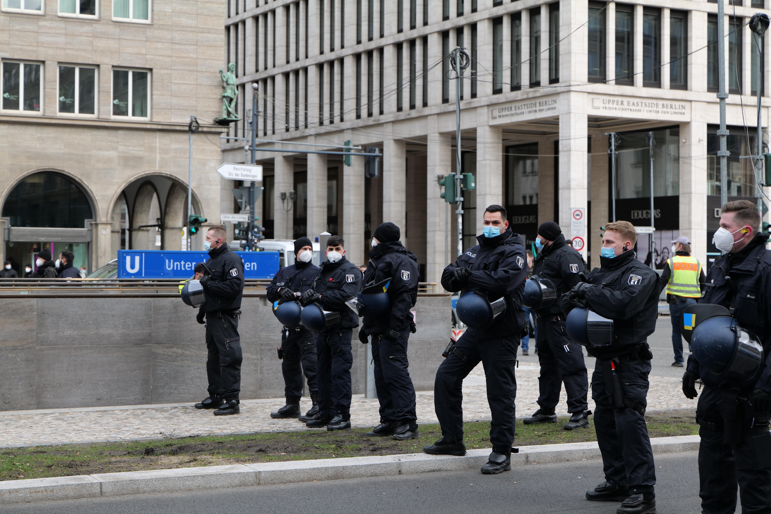 Gruppe von Polizisten in schwarzen Uniformen und Masken vor einem Berliner Gebäude mit Glasfenstern, Säulen und umliegenden städtischen Elementen.