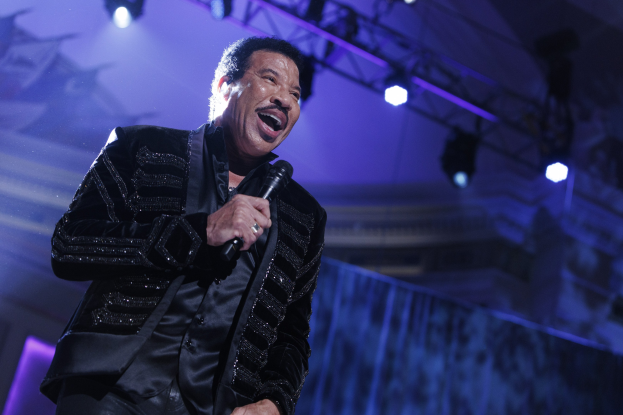 Lionel Richie bei den Grammys performend, in einem schwarzen Kleid und mit Mikrofon, mit hellen Bühnenlichtern, die eine dramatische Atmosphäre schaffen.