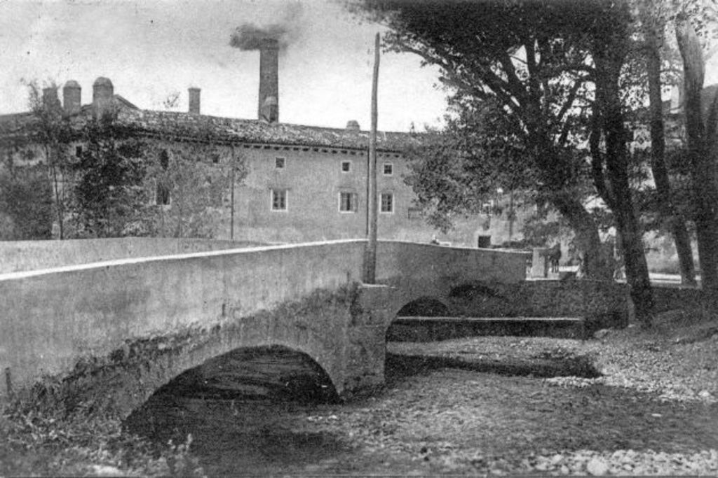Eine Szene mit Gebäuden und Bäumen, einem Pfahl und einer Brücke im Vordergrund, Wasser unten, Himmel oben und Rauch, der aus einem Schornstein aufsteigt.