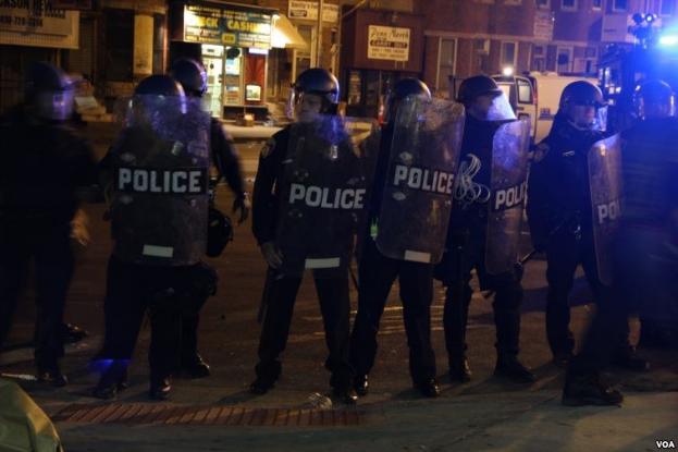 Gruppe von Polizisten in Helmen und Schilden auf einer Straße bei Nacht mit Gebäuden, Fahrzeugen und Laternen im Hintergrund.