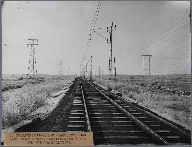 Schwarzes und weißes Foto einer Bahnschiene mit Strommasten und -drähten daneben, umgeben von Gras, mit Text unten.