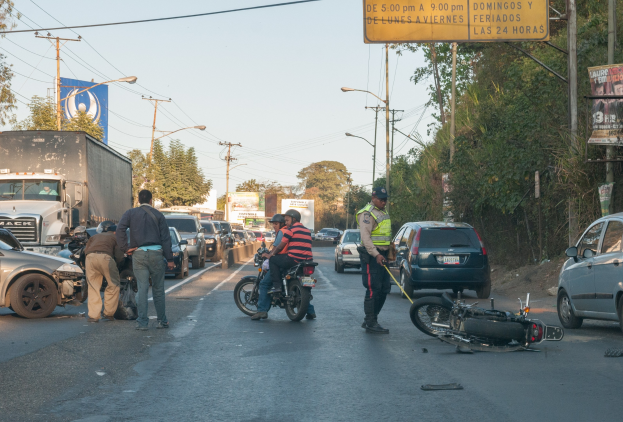 Eine Gruppe von Menschen steht um ein verunglücktes Motorrad am Stra├čenrand mit mehreren Fahrzeugen, darunter ein Lastwagen, und Hintergrundelementen wie B├Ąumen, Masten, Lichtern, Schildern und dem Himmel.