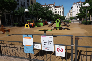 Ein umzäunter Stadtplatz-Spielplatz mit mehreren Schildern am Zaun, umgeben von Bäumen, Laternen, Gebäuden mit Fenstern und einem sichtbaren Himmel, mit verstreuten Objekten im Hintergrund.