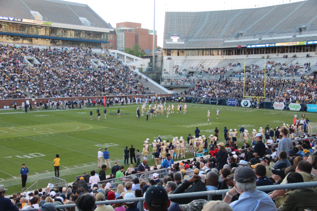 Ein Fussballspiel im Gange mit Spielern auf dem Feld und Zuschauern in den Rängen unter einem klaren blauen Himmel.