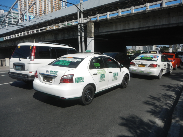 Eine Gruppe von Taxis entlang einer Straße mit einer Brücke, Strommasten, Gebäuden, Bäumen und einem klaren blauen Himmel im Hintergrund.