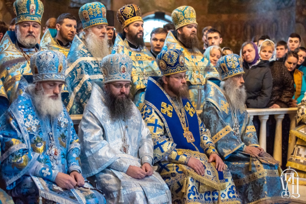 Eine Gruppe von Priestern in blauen und goldenen Gewändern sitzt vor einer Menge in einer Kirche mit braunen und cremefarbenen Wänden, getrennt durch ein Geländer.