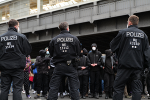 Polizisten in schwarzen Uniformen und Masken stehen vor einer Menge, mit einer Brücke und einem Gebäude im Hintergrund.