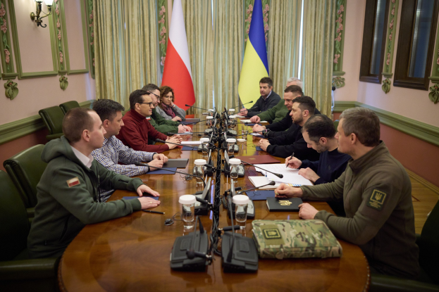 Gruppe von formell gekleideten Männern in einer Besprechung um einen Holztisch mit Mikrofonen, Dokumenten und Gläsern in einem Raum mit Flaggen, Vorhängen und an der Wand montierten Lampen.