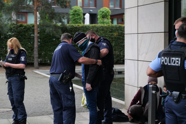 Eine Gruppe von Polizeibeamten steht vor einem Gebäude, eine Frau hält ein Buch und einen Stift in der Hand, während Menschen in der Nähe sitzen und stehen, mit Bäumen und Gebäuden im Hintergrund.
