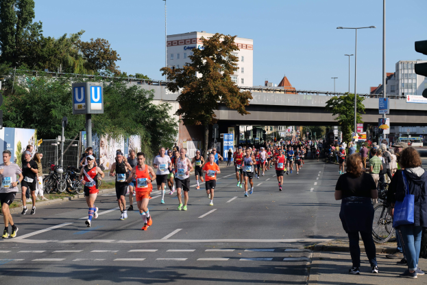 Gruppe von Menschen, die bei einem Marathon auf einer baumgesäumten Straße mit Laternen, Schildern, Fahrrädern, einem Zaun, Gras, einer Brücke, Gebäuden mit Fenstern und einem klaren blauen Himmel laufen.