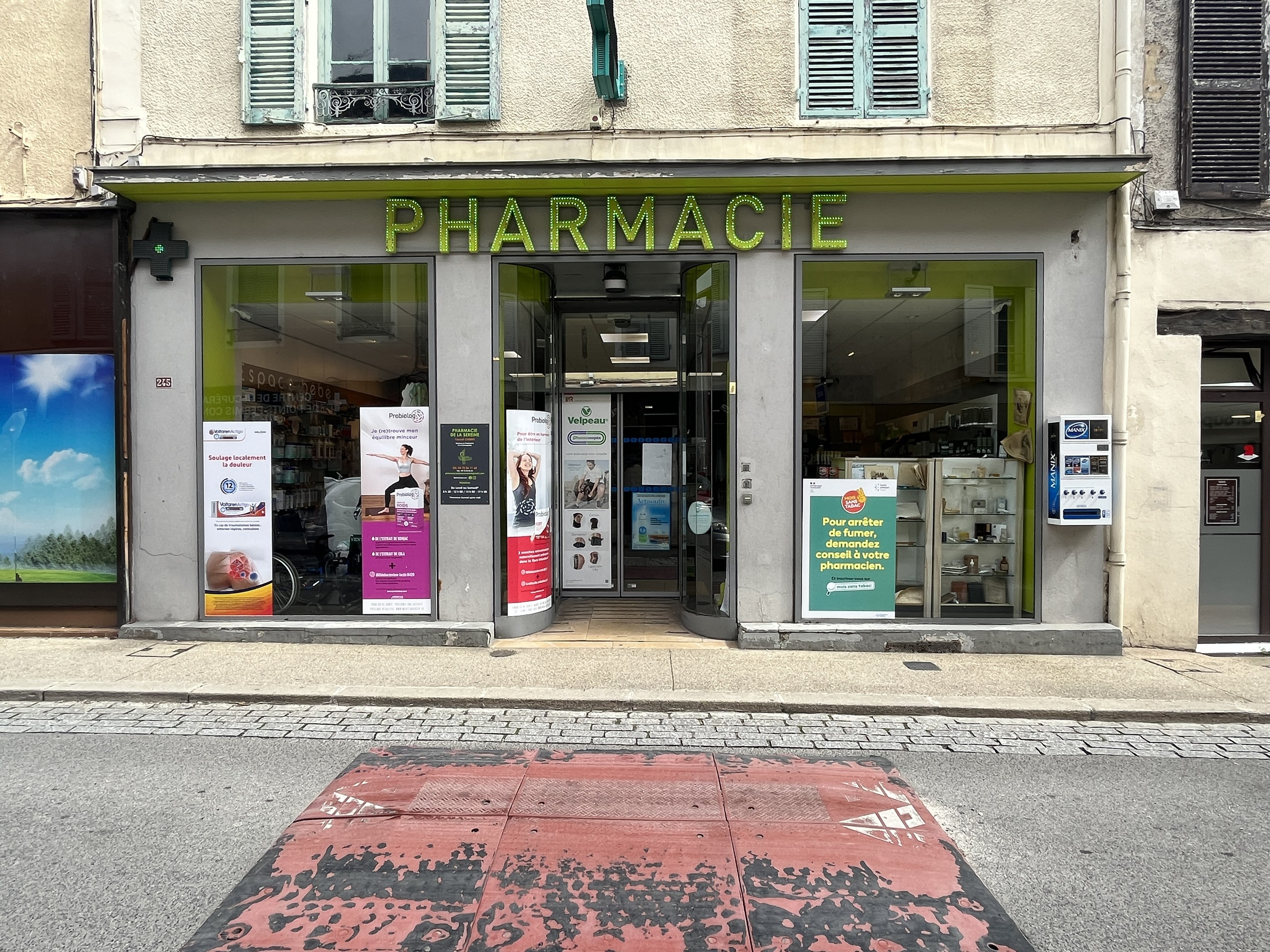 Apotheke mit Glaswänden und -türen, Plakate an den Innenwänden, an einer Straßenkreuzung gelegen.