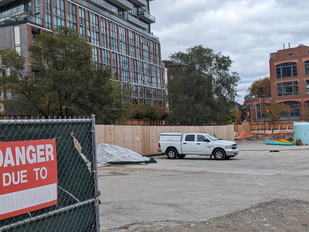 Truck parked near a building with a "Danger Due to Open Excavation" sign, trees and buildings in the background under a clear blue sky.