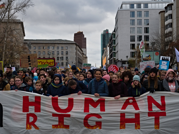 Eine Gruppe von Menschen mit einem "Menschenrechte"-Schild vor einer Menge mit Schildern und Fahnen, mit Bäumen, Gebäuden und Laternen im Hintergrund.