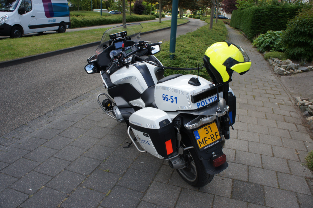 Ein Polizeimotorrad am Straßenrand geparkt mit einem Helm darauf, umgeben von Bäumen, Pfosten, Gebäuden, Pflanzen, Gras, Steinen und anderen Fahrzeugen im Hintergrund.