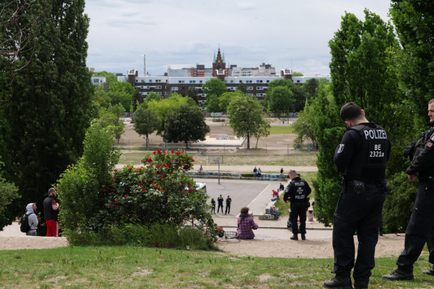 Zwei Polizeibeamte vor einer Gruppe von Menschen in einem Park mit Grünflächen, bunten Blumen und Gebäuden im Hintergrund unter einem klaren blauen Himmel.