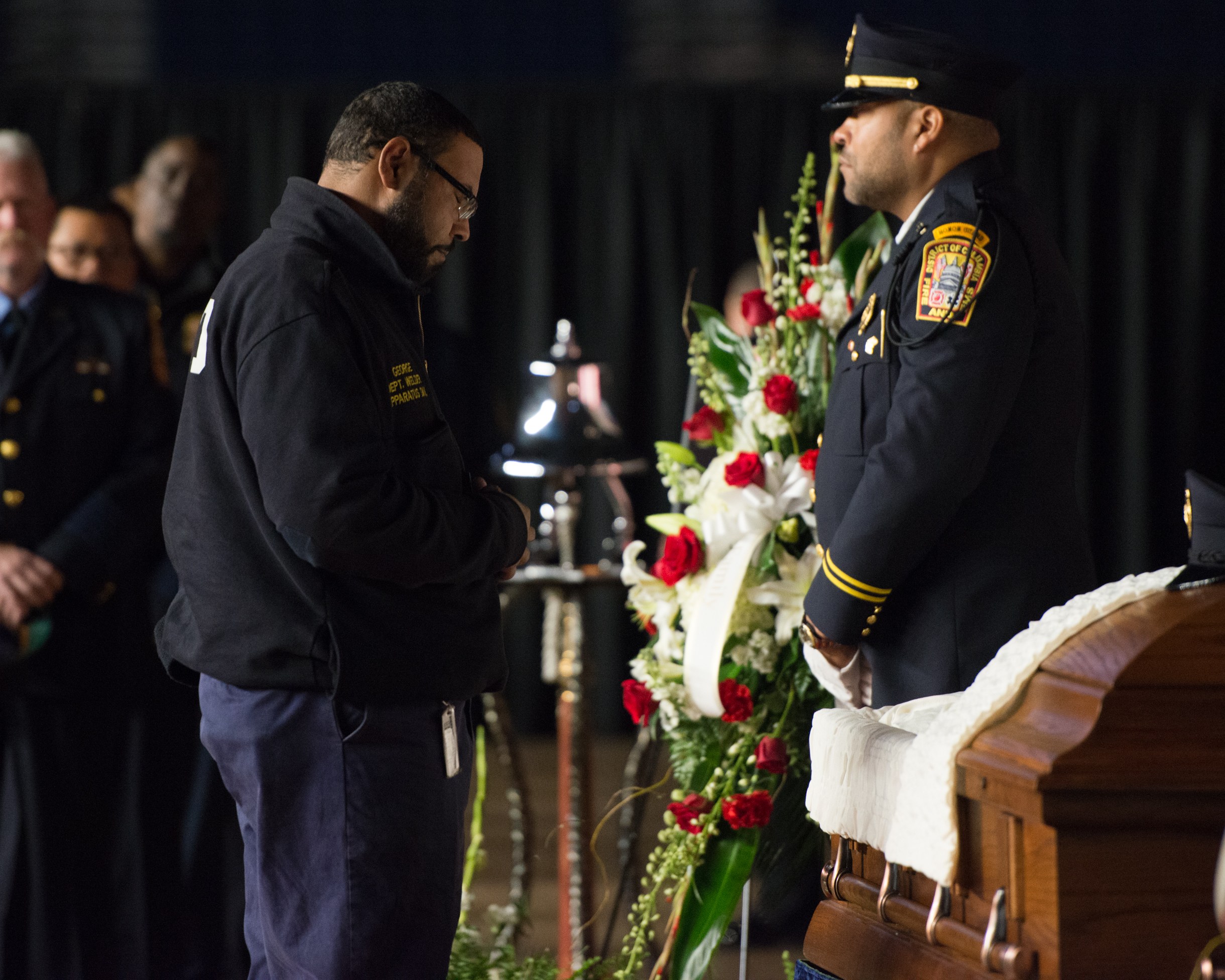 Zwei Polizisten in Uniform stehen feierlich neben einem Blumen geschmückten Sarg, mit einigen Personen im Hintergrund.